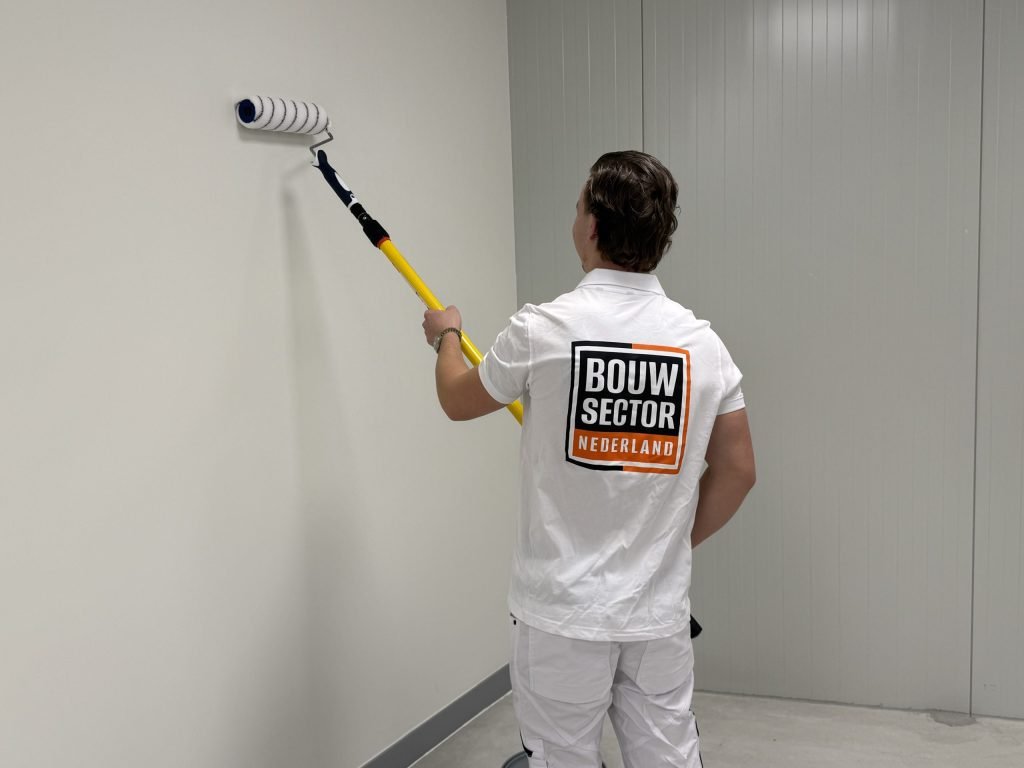 Schilder in witte werkkleding zet de roller aan een gele steel hoog op een lichte muur